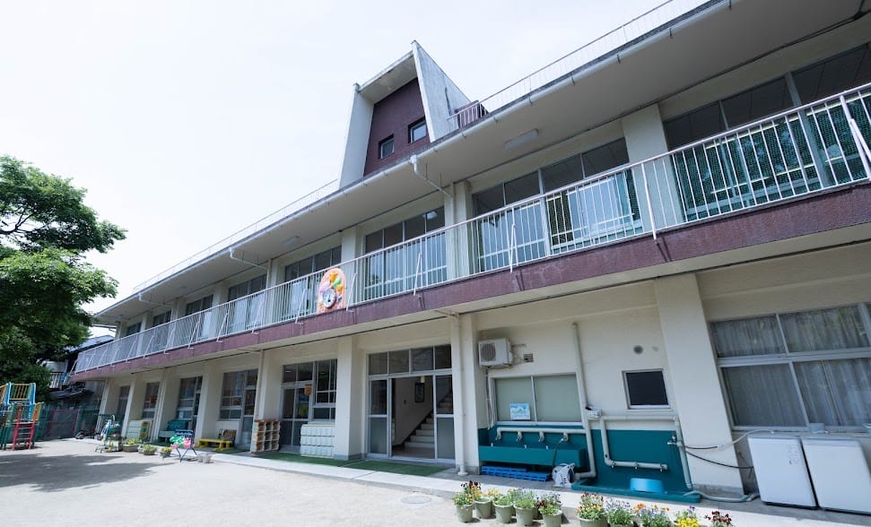 about_main | 保育士求人|大阪・兵庫・関西の保育士・幼稚園教諭の就職転職支援|保育のせかい Two-story building with balconies and a small outdoor play area to the left; potted plants line the front.