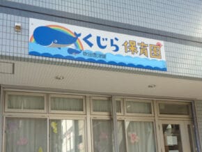 converted_v1-193c5720-8ad9-48b1-a2c5-97502d8d656c | 保育士求人|大阪・兵庫・関西の保育士・幼稚園教諭の就職転職支援|保育のせかい Front of a tiled building with a whale-themed sign くじら保育園 above glass doors.
