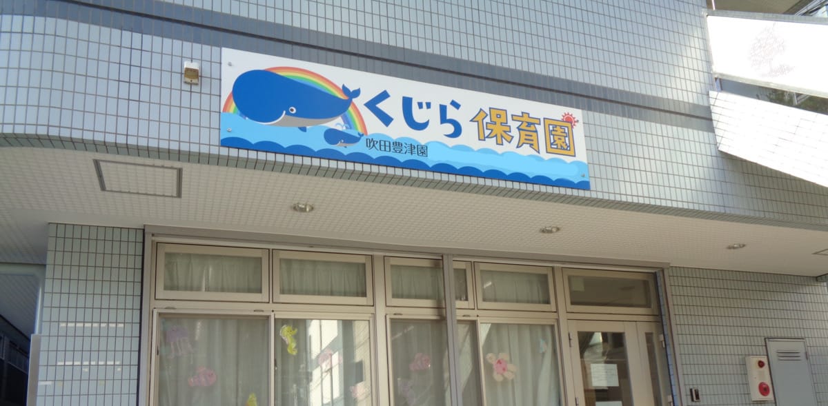 converted_v1-193c5720-8ad9-48b1-a2c5-97502d8d656c | 保育士求人|大阪・兵庫・関西の保育士・幼稚園教諭の就職転職支援|保育のせかい Front of a tiled building with a whale-themed sign くじら保育園 above glass doors.