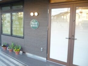 Exterior of a modern building entrance with glass doors, a circular sign on the gray wall, and potted plants outside.