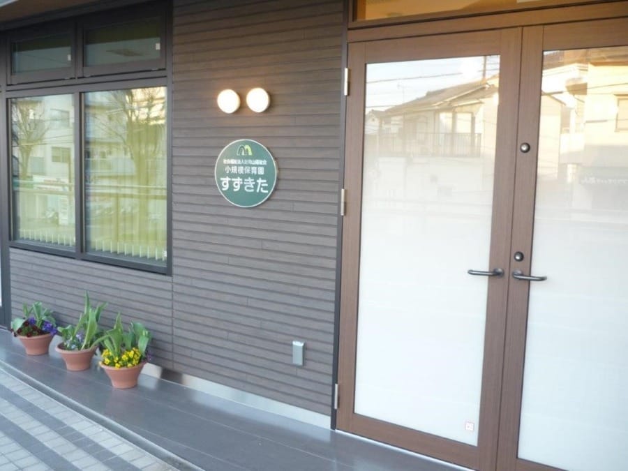 Exterior of a modern building entrance with glass doors, a circular sign on the gray wall, and potted plants outside.