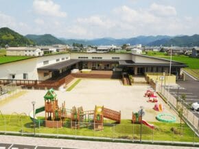 School playground with wooden play structures and slides, facing a large building and distant green hills.