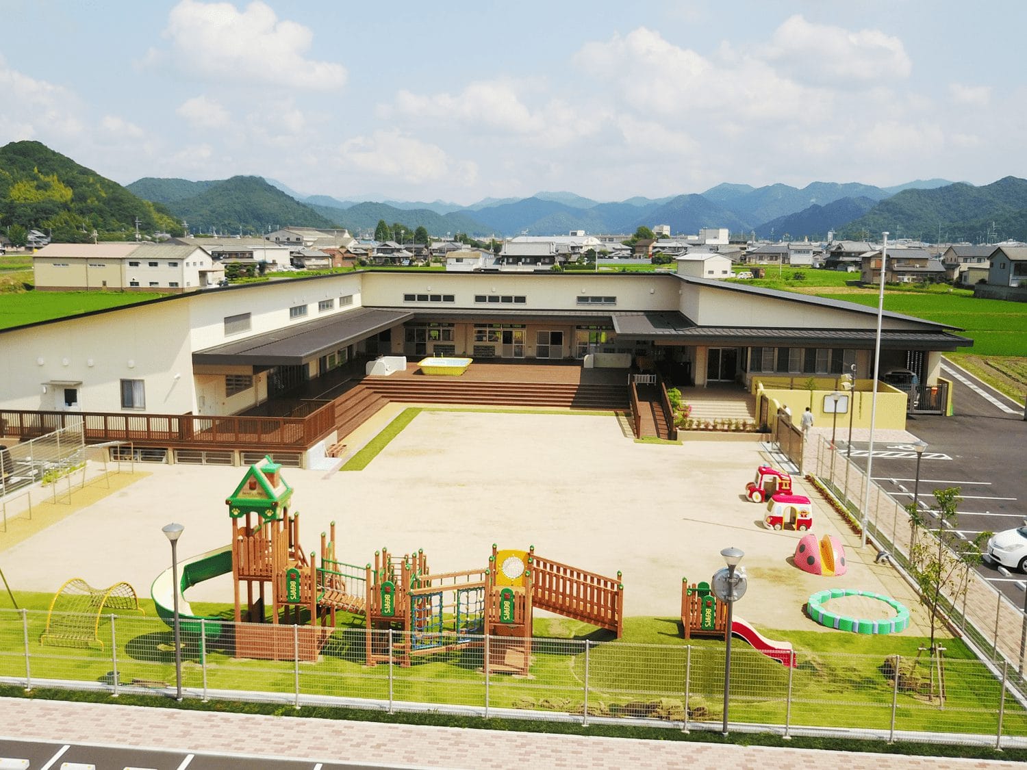 shisetsu_28 | 保育士求人|大阪・兵庫・関西の保育士・幼稚園教諭の就職転職支援|保育のせかい School playground with wooden play structures and slides, facing a large building and distant green hills.