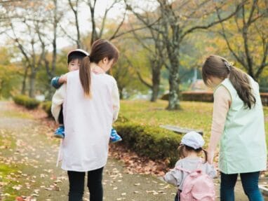 Two women walk with young children in a park during autumn; leaves on the path.