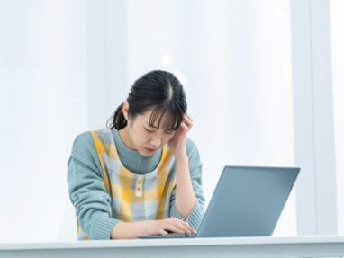 Young woman sits at a desk typing on a laptop, her hand on her temple as she concentrates.