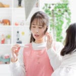 Woman in a pink apron holds a thermometer in one hand and talks on a smartphone in a bright, tidy room with shelves in the background.