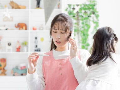 Woman in a pink apron holds a thermometer in one hand and talks on a smartphone in a bright, tidy room with shelves in the background.