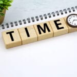 Wooden blocks spell 'TIME' with a small round clock at the end on a white desk, plus a plant, notebook, and pen nearby.
