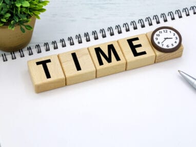 Wooden blocks spell 'TIME' with a small round clock at the end on a white desk, plus a plant, notebook, and pen nearby.