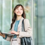 Professional woman in a beige blazer uses a laptop outdoors near glass office walls.