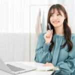 Young woman in a teal blouse sits at a bright desk with a laptop and notebook, holding a pen and smiling.