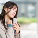 Young woman in a beige blazer holds a smartphone close to her chest, looking contemplative outside a modern building.