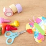 Colorful craft supplies on a wooden table: scissors, tape rolls, and bright plastic craft pieces with a painted paper flower.