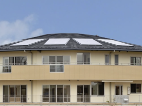 Modern two-story yellow house with a curved balcony and solar panels on the sloped roof under a blue sky.