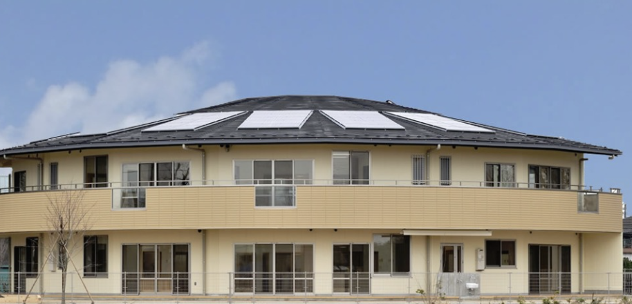 Modern two-story yellow house with a curved balcony and solar panels on the sloped roof under a blue sky.