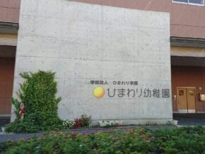Concrete wall with a sign for 'ひまわり幼稚園' (Himawari Kindergarten), yellow sun logo, and an entrance to a brick building on the right, with plants in front.