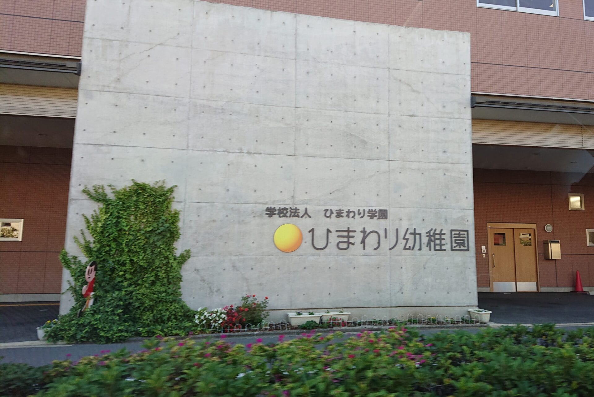 Concrete wall with a sign for 'ひまわり幼稚園' (Himawari Kindergarten), yellow sun logo, and an entrance to a brick building on the right, with plants in front.