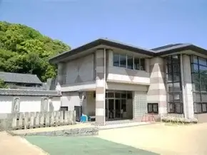 Modern two‑story building with large glass windows, concrete walls, and a green hill nearby on a sunny day