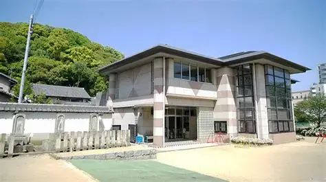OIP 8 | 保育士求人|大阪・兵庫・関西の保育士・幼稚園教諭の就職転職支援|保育のせかい Modern two‑story building with large glass windows, concrete walls, and a green hill nearby on a sunny day