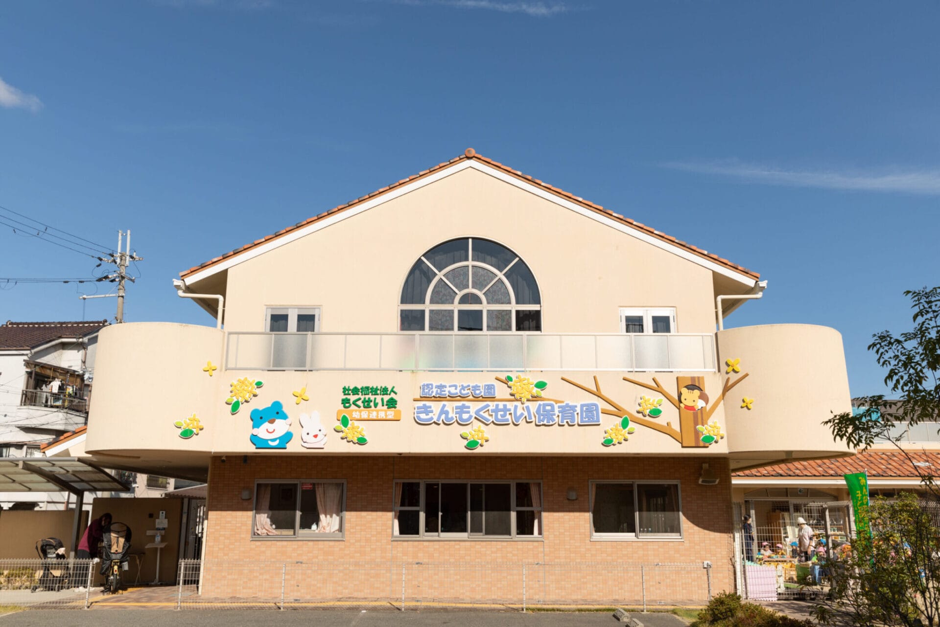 Front view of a beige daycare building with a large arched window, colorful cartoon characters, and a welcoming sign beneath a clear blue sky.