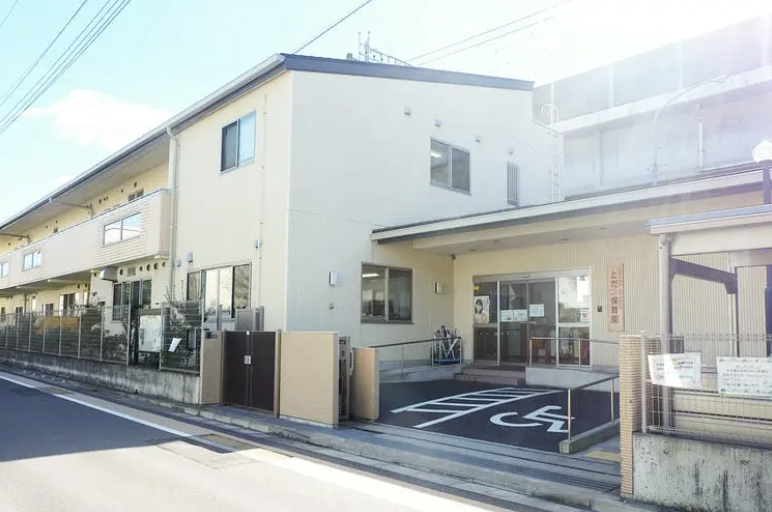 スクリーンショット 2026-09-11 145810 | 保育士求人|大阪・兵庫・関西の保育士・幼稚園教諭の就職転職支援|保育のせかい Exterior of a beige medical clinic with a marked disabled parking space near the entrance, along a street.