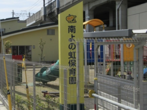 Entrance area of a Japanese nursery/kindergarten with a tall yellow sign and a fenced playground featuring colorful play structures in the background.