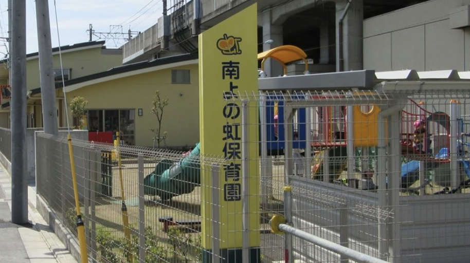 Entrance area of a Japanese nursery/kindergarten with a tall yellow sign and a fenced playground featuring colorful play structures in the background.