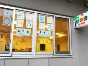 Daycare storefront with large windows spelling OHANA in blue letters, Japanese characters, and a palm-tree sign reading OHANA on the right side outside.