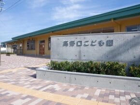 Exterior of a yellow school building with a gray sign bearing Japanese characters in front and a patterned brick walkway.”,“Entrance area of a childcare center with a green roof, concrete sign, and shrub bed in front.”,“Front view of a school/care center with a concrete sign reading 高野口こども園 and manicured greenery.”,“Wide shot of a childcare facility entrance under a bright blue sky, showing signage and a tiled plaza.”,“Concrete sign with Japanese text beside a row of shrubs at the front of a yellow building.”]}}lang_iso:en-US} Let's correct formatting. Ensure valid JSON. The assistant included stray characters. Let's produce proper JSON. The instruction expects {