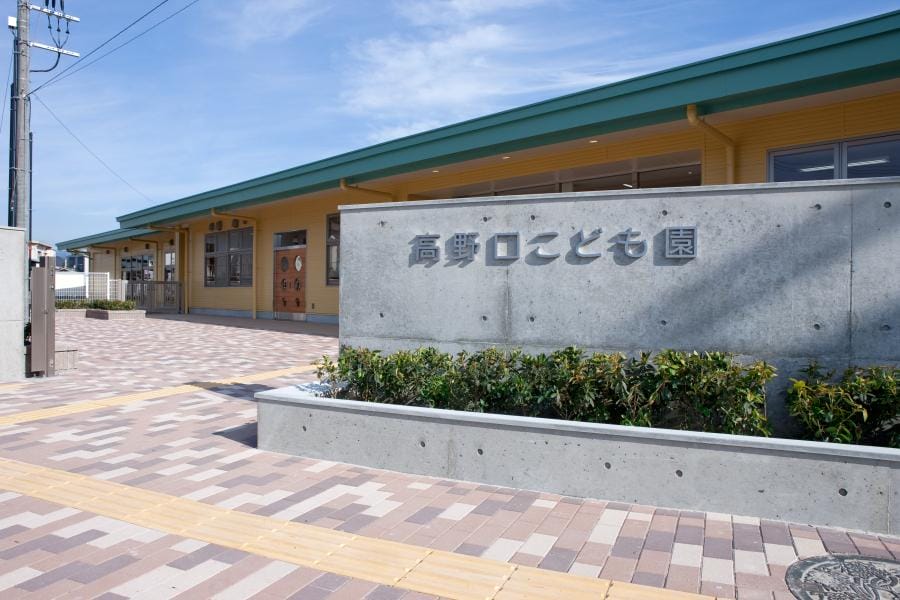 Exterior of a yellow school building with a gray sign bearing Japanese characters in front and a patterned brick walkway.”,“Entrance area of a childcare center with a green roof, concrete sign, and shrub bed in front.”,“Front view of a school/care center with a concrete sign reading 高野口こども園 and manicured greenery.”,“Wide shot of a childcare facility entrance under a bright blue sky, showing signage and a tiled plaza.”,“Concrete sign with Japanese text beside a row of shrubs at the front of a yellow building.”]}}lang_iso:en-US} Let's correct formatting. Ensure valid JSON. The assistant included stray characters. Let's produce proper JSON. The instruction expects {