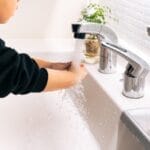 Child washing hands under running water at a bathroom sink with a plant nearby