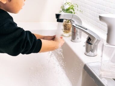 Child washing hands under running water at a bathroom sink with a plant nearby