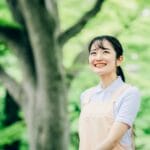 Smiling young woman in a light blue shirt and beige apron stands outdoors in a green park.