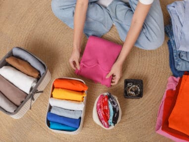 Top-down view of a person packing a suitcase with neatly folded clothes in colorful stacks on a woven rug