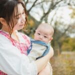 Mother holds a baby in a park with autumn trees in the background.