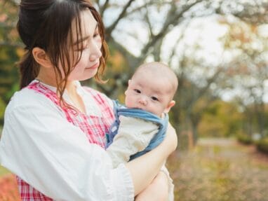 Mother holds a baby in a park with autumn trees in the background.