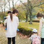 Two women walk with young children in a park during autumn leaves on the ground, facing away from the camera.
