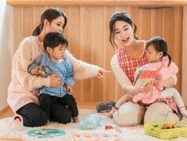 Two women with two small children play on a rug at home; one points while the other holds a tablet for the child in a pink dress.