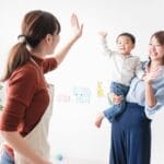 Two women share a joyful high-five while a mother holds a smiling child in a bright, playful room with animal decals on the wall.