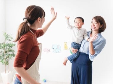 Two women share a joyful high-five while a mother holds a smiling child in a bright, playful room with animal decals on the wall.