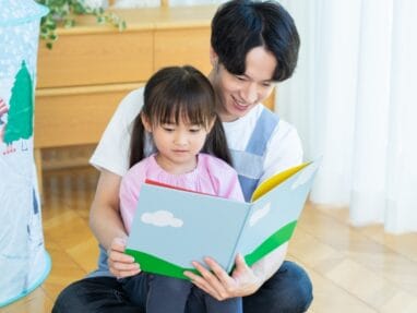 34596203_s | 保育士求人|大阪・兵庫・関西の保育士・幼稚園教諭の就職転職支援|保育のせかい Father and young daughter reading a colorful picture book together on a wooden floor.