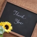 Chalkboard with 'Thank You' in cursive, accompanied by a bouquet of sunflowers and green hydrangeas on a wooden table.