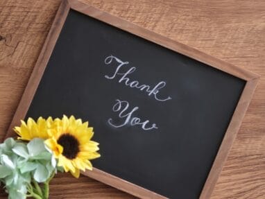 Chalkboard with 'Thank You' in cursive, accompanied by a bouquet of sunflowers and green hydrangeas on a wooden table.