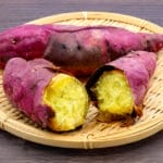 Purple-skinned sweet potatoes on a round bamboo tray, one cut open to show bright yellow flesh.