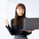 Woman in a black top holds a laptop and pumps her fist in celebration, smiling at the screen-satisfaction pose.