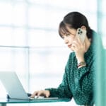 Woman in teal blouse sits at glass desk, talking on a smartphone while typing on a laptop.