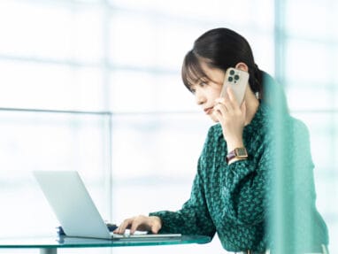 Woman in teal blouse sits at glass desk, talking on a smartphone while typing on a laptop.