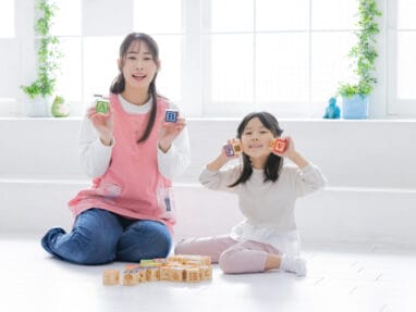 Mother and daughter sit on a bright white floor, holding alphabet blocks A and B and smiling at the camera with wooden blocks on the ground nearby.