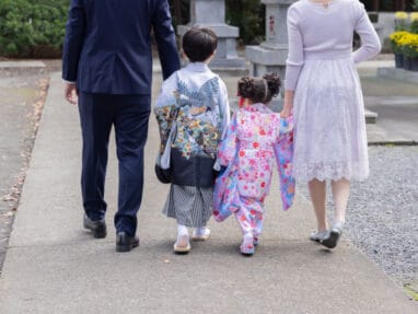 35368249_s | 保育士求人|大阪・兵庫・関西の保育士・幼稚園教諭の就職転職支援|保育のせかい Family walking away along a temple path: man in dark suit, woman in lilac dress, and two children in colorful kimonos holding hands as they stroll toward the shrine area.