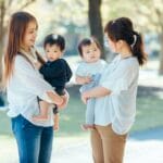 Two women in white tops hold young children and chat in a sunny park, smiling at each other.